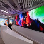 "Interior view of Design 710 Atlantic City Dispensary showing a curved hallway with light wood flooring and white walls. The space features an industrial-style ceiling with exposed white linear slats and integrated lighting. A large curved digital display screen dominates the right wall, showing vibrant abstract imagery in red, blue, and green colors with "ATLANTIC CITY" text overlay. In the background, vintage travel posters are visible on the left wall, including Atlantic City promotional artwork. The modern dispensary design combines sleek architectural elements with dynamic digital displays and nostalgic tourism imagery."