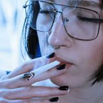 A person with short dark hair, glasses, and black nail polish is smoking a cigarette—perhaps rolled with some local weed—with their eyes closed as smoke drifts around their face.