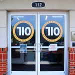 Entrance of a dispensary with glass double doors labeled “Dispensary 10 Design Atlantic City.” Signs on the doors, a doorbell to the right, and a black oval address number “112” above the entrance make it stand out among dispensaries near me.