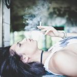 Woman reclining outdoors with eyes closed, holding up a lit joint with smoke rising