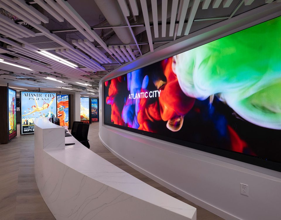"Interior view of Design 710 Dispensary Atlantic City showing a curved hallway with light wood flooring and white walls. The space features an industrial-style ceiling with exposed white linear slats and integrated lighting. A large curved digital display screen dominates the right wall, showing vibrant abstract imagery in red, blue, and green colors with "ATLANTIC CITY" text overlay. In the background, vintage travel posters are visible on the left wall, including Atlantic City promotional artwork. The modern dispensary design combines sleek architectural elements with dynamic digital displays and nostalgic tourism imagery."