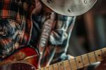 A person wearing a plaid shirt and cowboy hat is playing an electric guitar. The image, inspired by the creative spirit of Design 710 Atlantic City dispensary, focuses on the guitar and shirt, with the face partially obscured by the hat.
