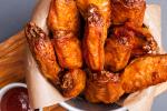 A bowl lined with parchment paper filled with crispy, golden-brown chicken wings, with a small dish of red dipping sauce on the side.