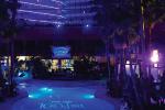 A dimly lit indoor space with blue and purple lighting, palm trees, and a bar area in the background. The words “Key West” are projected onto the floor.