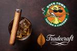 A glass of whiskey with ice and a cigar beside logos for Duck Town Tavern & Liquor, featuring a cool duck, and Tradecraft, with a brown leaf and elegant script text.