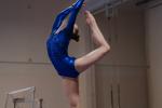 A gymnast in a shiny blue leotard performs a highly flexible backbend, holding one leg overhead while standing on the other leg in an indoor gym.