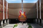 A large guitar-shaped Hard Rock Cafe sign stands above a driveway entrance between two modern multi-story buildings under a partly cloudy blue sky.