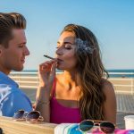 A man and woman sit on a bench at the beach. The woman is smoking and exhaling smoke, while the man looks at her. Sunglasses and drinks are on the bench, with the ocean and blue sky in the background.