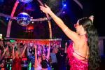 A performer in a pink costume stands on stage with arms raised, facing a lively crowd dancing and cheering under colorful lights and a disco ball in a nightclub.