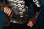 A person wearing detailed medieval-style armor grips the handle of a sword at their waist. The armor features metal plates and leather straps, with the individuals hands partially covered by red cloth.