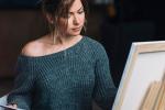 A woman wearing a gray sweater sits and paints on a blank canvas, focused on her artwork in a softly lit room.