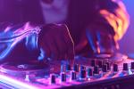 Close-up of a DJs hands adjusting controls on a mixing console, with colorful purple and blue lighting creating a lively atmosphere.