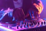 Close-up of a DJ’s hands adjusting controls on a mixing console, with purple and blue lighting creating a vibrant atmosphere.