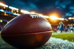A close-up of an American football lying on the field with a blurred stadium and players in the background under bright stadium lights.