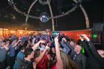 A large crowd of people are dancing and taking selfies at a lively indoor party, with colorful lights and a disco ball hanging from the ceiling.