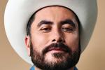 A man with a dark beard and mustache wears a white cowboy hat and a blue shirt, looking confidently at the camera against a neutral background.