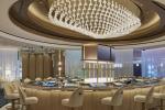 A modern, elegant restaurant interior features a large circular bar with cream-colored chairs and a striking, sculptural chandelier hanging above. The lighting is warm and the decor is sophisticated and upscale.