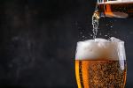 Close-up of a glass of beer being poured from a bottle, with golden liquid, bubbles, and a foamy head against a dark background.