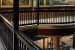 Interior view of a museum or gallery showing a dark wooden railing on an upper level, with framed paintings hanging on brick walls in the background. The space appears quiet and well-lit.