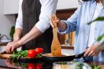 Two people are cooking together in a kitchen. One is stirring food in a frying pan with a wooden spatula, while the other is preparing fresh vegetables, including tomatoes and leafy greens, on a counter.