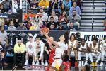 A basketball player in a white and yellow uniform leaps toward the hoop for a layup as a defender in red attempts to block him; spectators and teammates watch from the stands and bench.