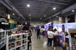 A busy convention hall with people walking and browsing various vendor booths filled with merchandise under bright lights. Shelves display colorful products on the left, and banners decorate the booths on the right.