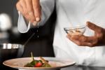 A chef in a white coat sprinkles seasoning from a small glass bowl onto a plated dish with vegetables, garnishing it carefully.