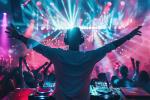 A DJ wearing headphones stands with arms raised behind a mixing console, facing a lively crowd dancing under colorful, vibrant lights in a packed nightclub.