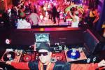 A DJ wearing sunglasses and a hat stands behind turntables and mixers in a vibrant nightclub, with colorful lights and people dancing on the crowded dance floor in the background.