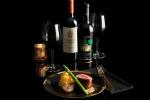 A gourmet meal of steak and potatoes is plated with asparagus, accompanied by two glasses of red wine, two wine bottles, and a gold candle holder, all set against a black background.