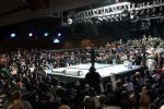 A large crowd fills an indoor arena around a brightly lit wrestling ring, with spectators seated on all sides and spotlights illuminating the ring area.