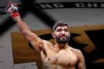 A male MMA fighter with a beard raises his right arm in victory inside a cage, wearing red gloves. The background shows part of a fighting championship logo.