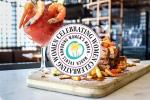 A shrimp cocktail and a dish of fries with meat are served on a tray. A circular Celebrating Women logo for the Bahamas Women’s Food Festival is displayed in the foreground.
