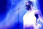 A singer passionately performs on stage, wearing a white tank top and illuminated by dramatic blue and purple stage lights, with a microphone stand in the background.