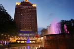 A tall, illuminated hotel building stands behind a park with a fountain lit in purple and pink at dusk. Festive lights and a decorated Christmas tree add color to the lively evening scene.