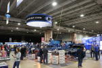 An indoor trade show with people walking around and visiting booths; large blue farm equipment is on display under a hanging sign that reads NEW HOLLAND. Bright lights illuminate the spacious hall.