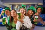 Five people dressed in green St. Patrick’s Day costumes smile and hold glasses of green beer in a festive bar setting, celebrating together.