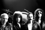 Four men in black leather jackets stand in front of a drum set with a dark background. They have serious expressions and distinctive 1970s–80s hairstyles, including long curly and wavy hair, and one has a prominent mustache.