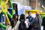 Four people talk to a man at a booth with inflatable structures and informational materials at an indoor event or expo.