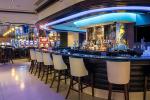 Modern bar with a curved counter, high-backed white stools, illuminated shelves displaying bottles, and blue accent lighting. The atmosphere is stylish and inviting, with additional seating and soft lighting in the background.