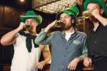 Three men wearing green hats celebrate in a bar, each drinking a pint of beer. The atmosphere is festive, and they appear to be enjoying St. Patrick’s Day.