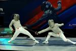 Two fencers in protective gear duel with swords on a lit platform; one lunges forward with a sword while the other leans back to evade, against a backdrop featuring a large image of another fencer.