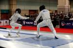 Two fencers in white protective gear and masks compete on a marked strip in an indoor arena, with a red carpet and spectators watching in the background.