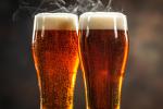 Two tall glasses filled with amber beer, topped with frothy white foam. The beer is cold, with condensation on the glasses, and a bit of steam rises above them. The background is blurred and dark.
