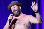 A bearded man wearing a hat and no shirt holds a microphone and gestures with his hand while performing on stage with purple lighting in the background.