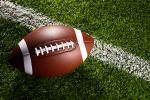 A brown American football with white laces rests on green grass, positioned next to a white yard line on a football field.