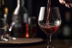 A close-up of red wine being poured into a glass, with blurred bottles and a warm, dimly lit background.