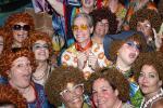 A group of people wearing curly red wigs, colorful retro clothing, and large glasses, smiling and posing closely together at a lively, festive event.