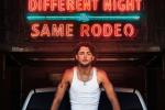 A man in a white tank top sits in front of a neon sign that reads Different Night Same Rodeo in bright red and blue letters.