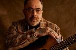 A man with glasses and a trimmed beard, wearing a plaid jacket, sits holding an acoustic guitar against a brown background.